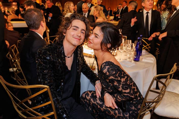 Two people in elegant evening attire pose at a formal event with a crowd in the background