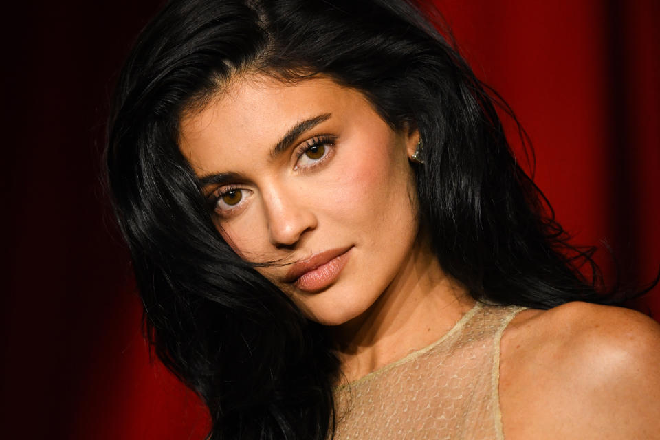 Kylie Jenner poses with sleek hairstyle and sheer textured top, set against a plain backdrop