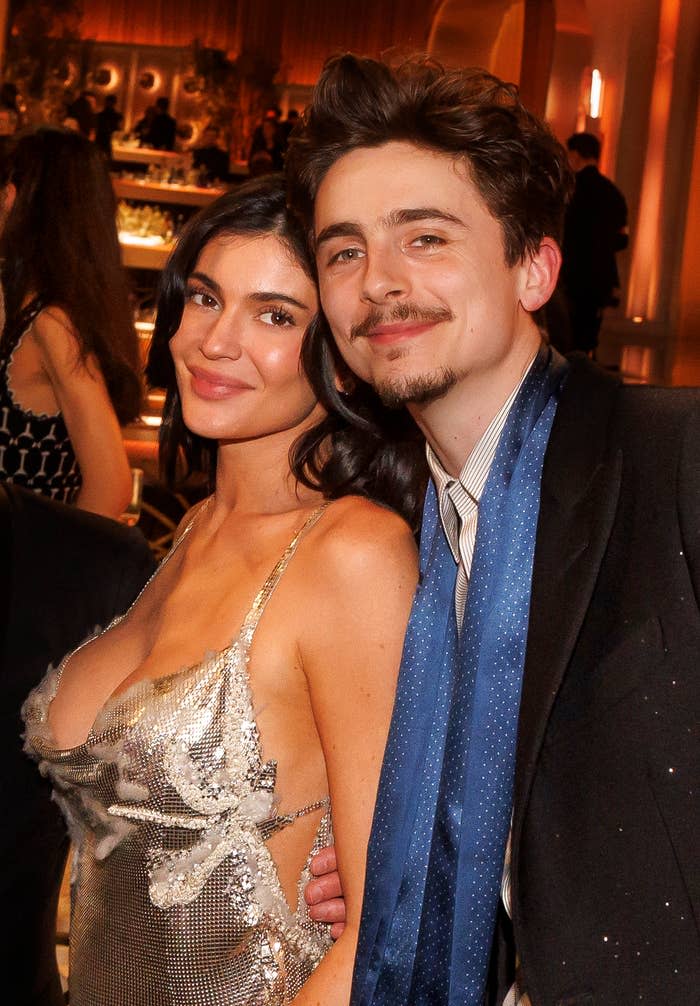 Two individuals at a formal event, one in a sequined dress and the other in a suit with a blue scarf, posing closely and smiling for the camera
