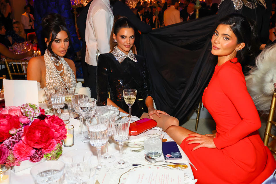 Kim Kardashian, Kendall Jenner and Kylie Jenner dressed glamorously sit at a formal dinner table, surrounded by elegant tableware and floral arrangements