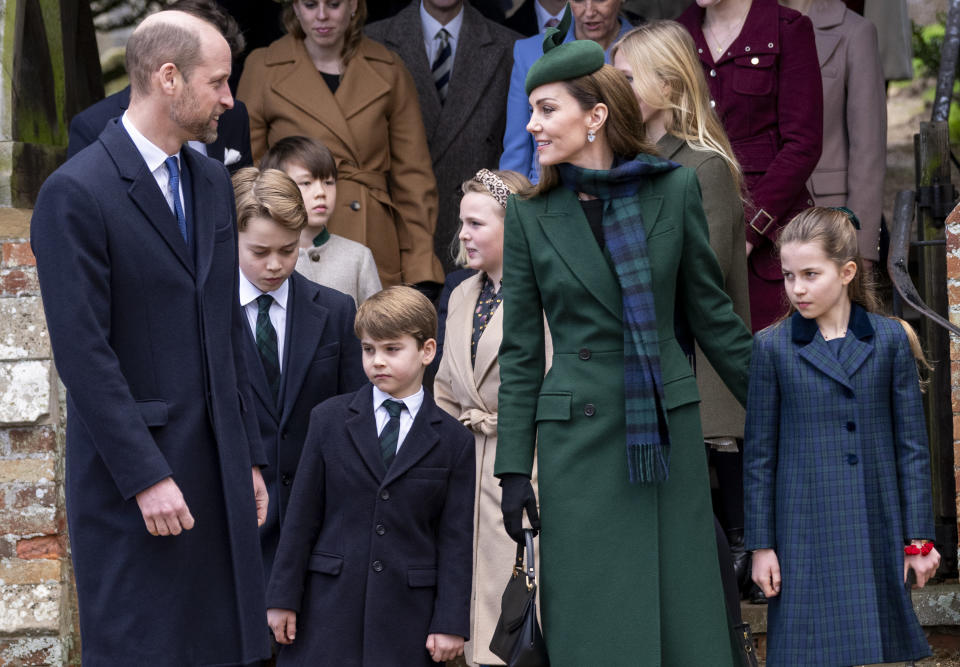 Prince William wearing a blue coat smiling and looking at Kate Middleton, in a green coat and plaid scarf, standing with Prince George, Princess Charlotte and Prince Louis outside church
