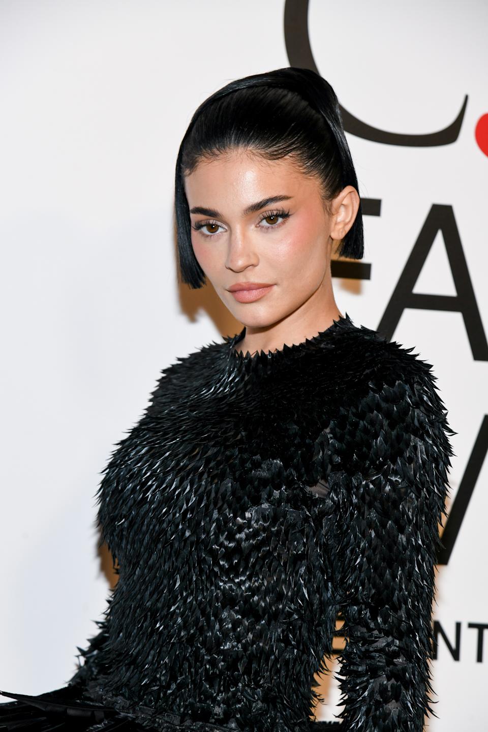Person in chic black textured outfit on red carpet, hair styled back, neutral expression