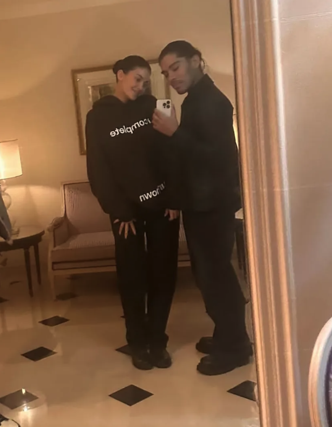 Two people pose in a mirror selfie. The person on the left wears a casual hoodie and pants. The person on the right wears a dark outfit
