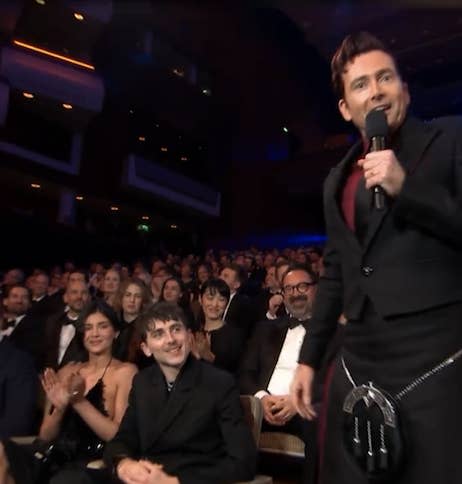 David Tennant on stage in a stylish suit speaking into a microphone, with Timothee Chalamet and Kylie Jenner clapping and smiling in support