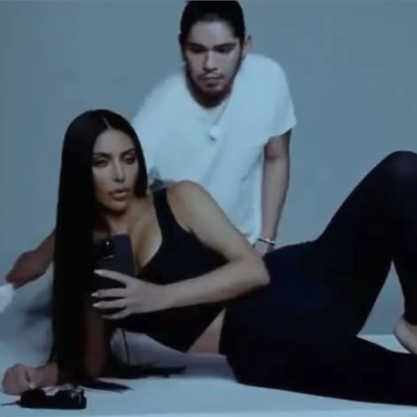 Person lounging with long hair, wearing sleek, fitted attire. Another person in a casual tee in the background, possibly in a studio setting