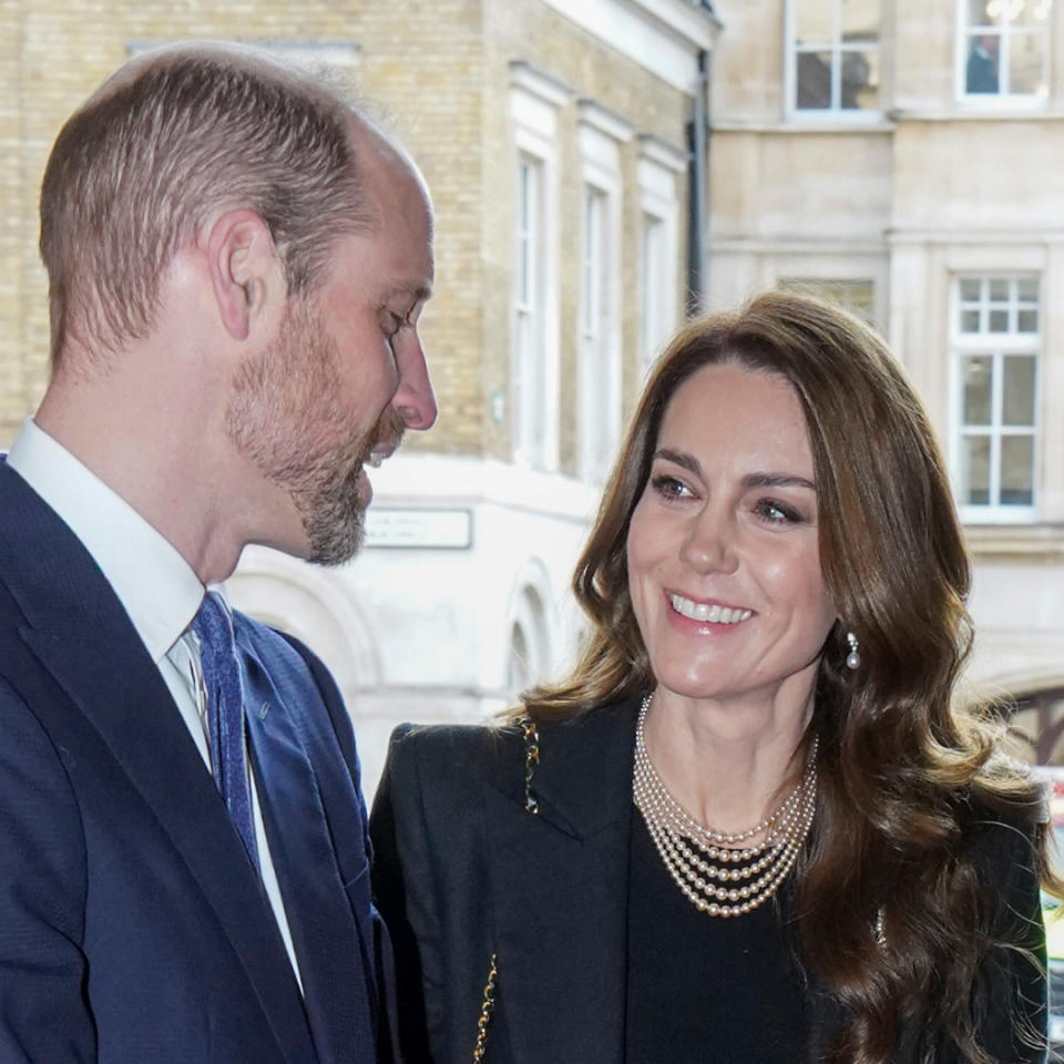 Kate Middleton has long, wavy brunette hair and wears a black jacket with a black top and a pearl necklace while gazing at husband Prince William's beard.