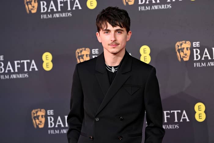 Timothée Chalamet in tailored dark suit on a BAFTA red carpet, standing against a backdrop with BAFTA logos