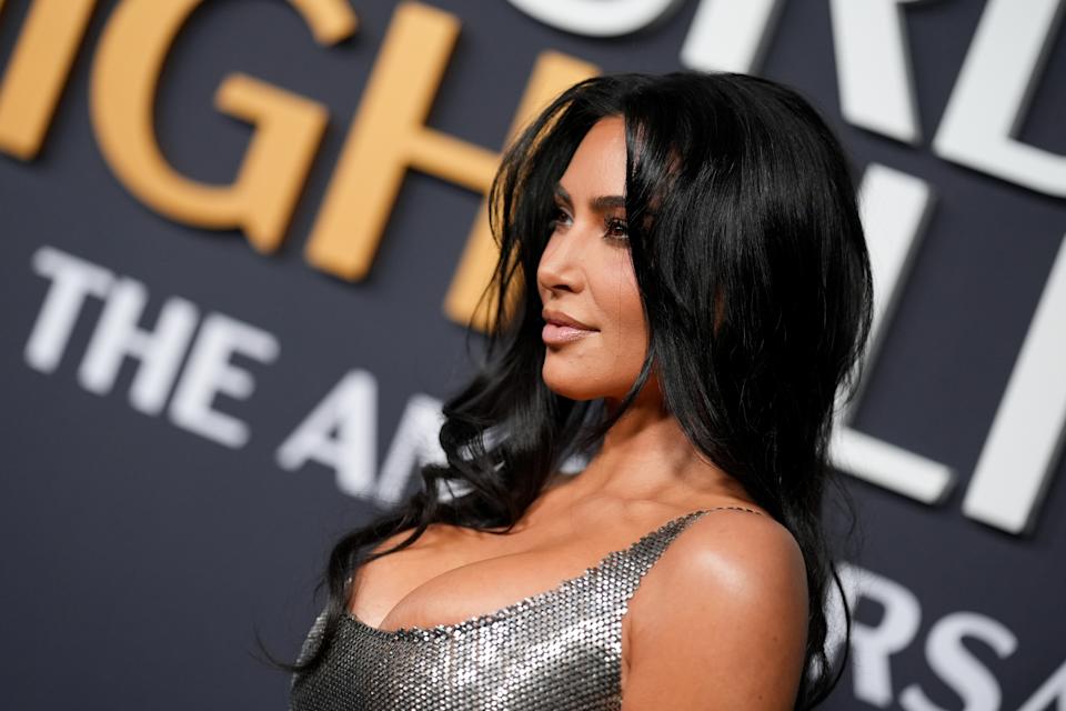 A celebrity poses on the red carpet in a shimmering, sleeveless dress with structured shoulders, against a backdrop with large text