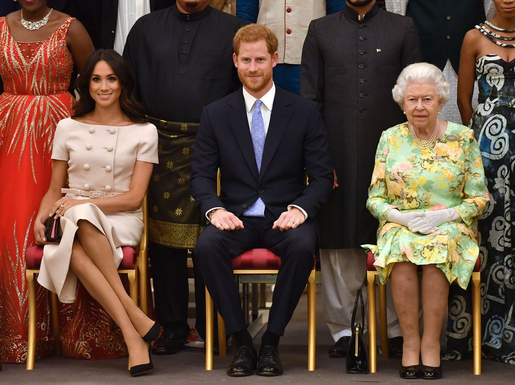 Meghan sitting in blush Prada outfit beside harry and queen elizabeth
