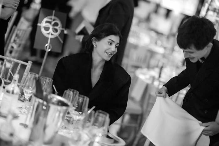 Person in formal attire sitting at a table, smiling at another person holding a napkin. Tables set for a formal event are in the background