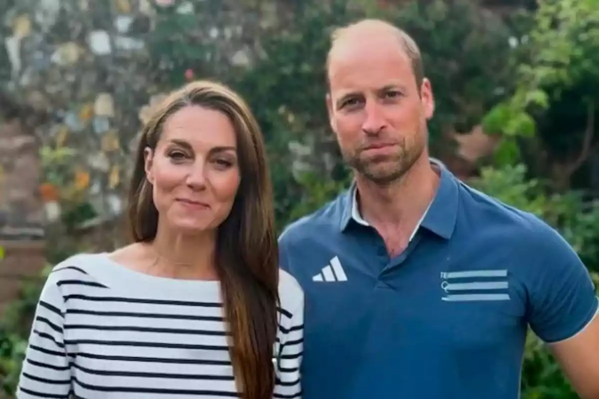 Kate Middleton in a striped T-shirt and Prince William in a blue T-shirt pose together outdoors against a backdrop of greenery and a stone wall.