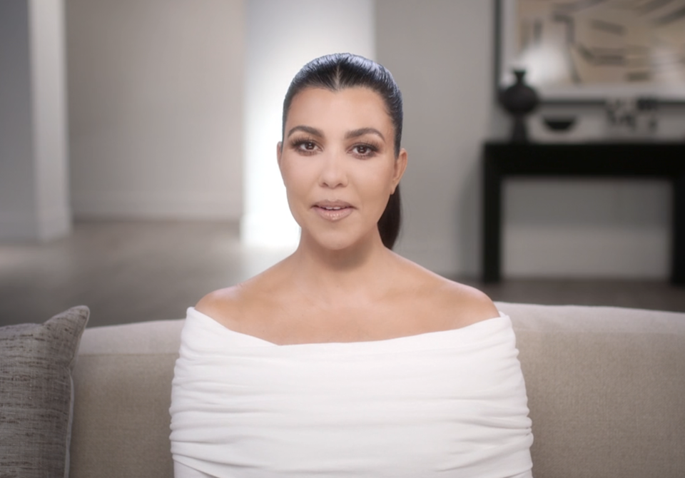 Kourtney Kardashian sitting on a couch in a modern living room, speaking directly to the camera