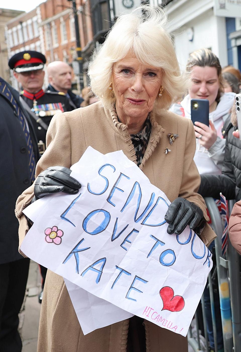 Queen Camilla holding a sign for Kate Middleton outdoors surrounded by people.