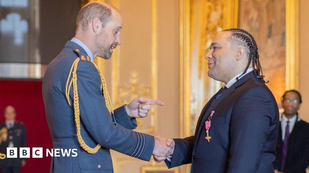 Diana Award campaigner collects OBE from Prince of Wales Alex Holmes shaking the hand of Prince William, the Prince of Wales, in a room in Windsor Castle. Prince William is wearing an army uniform, and is pointing at Alex. Alex is wearing a blue suit, with his OBE medal on his lapel. His hair is braided.