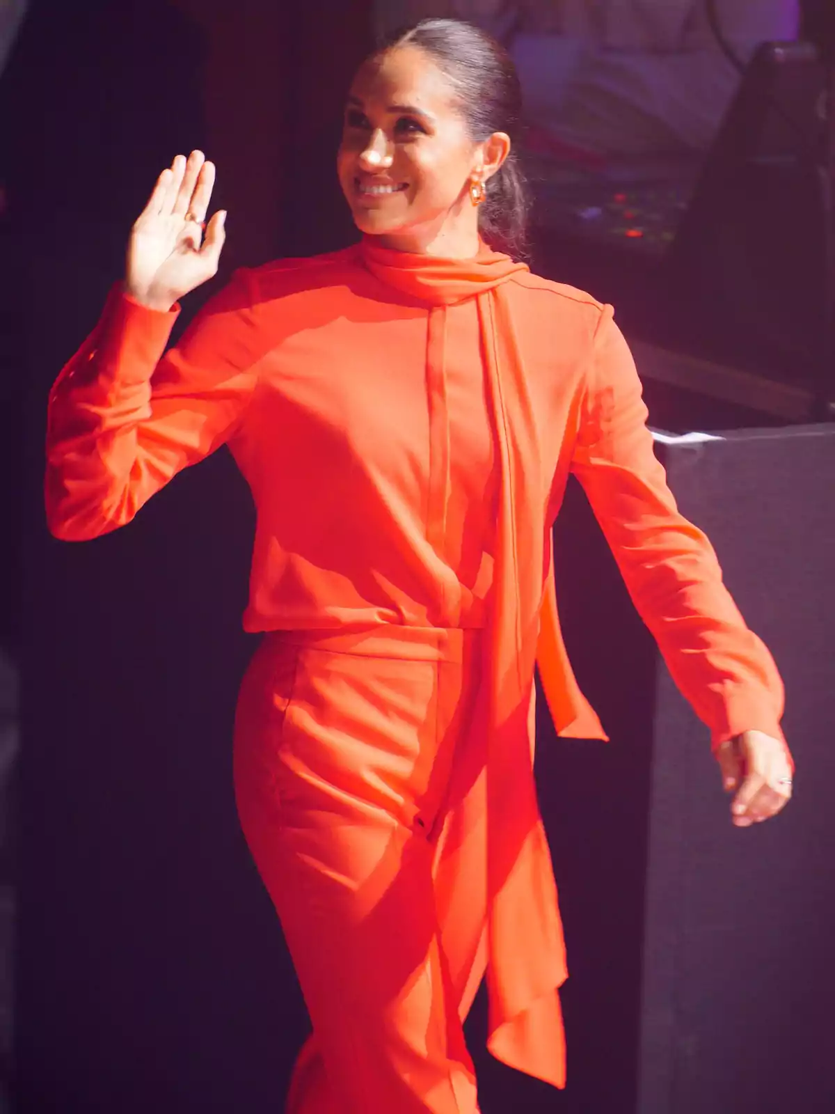 Meghan Markle, in a bright red outfit, smiles and waves as she walks.