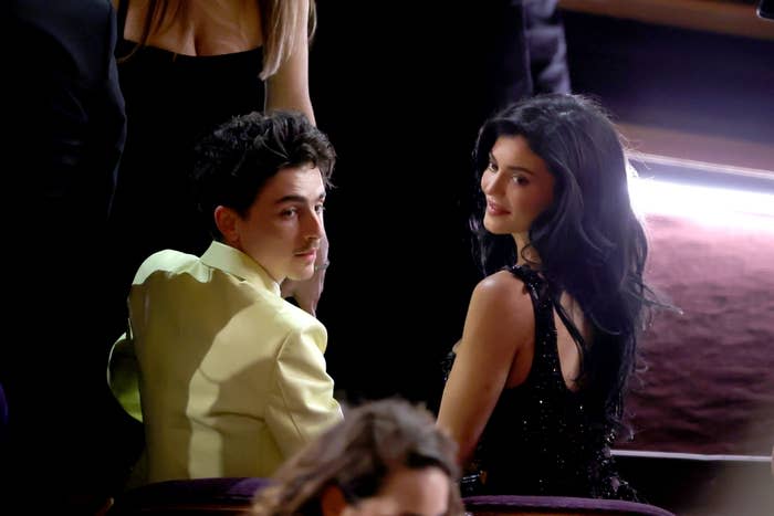 Timothée Chalamet and Kylie Jennerin formal attire seated at an event, looking over their shoulders toward the camera