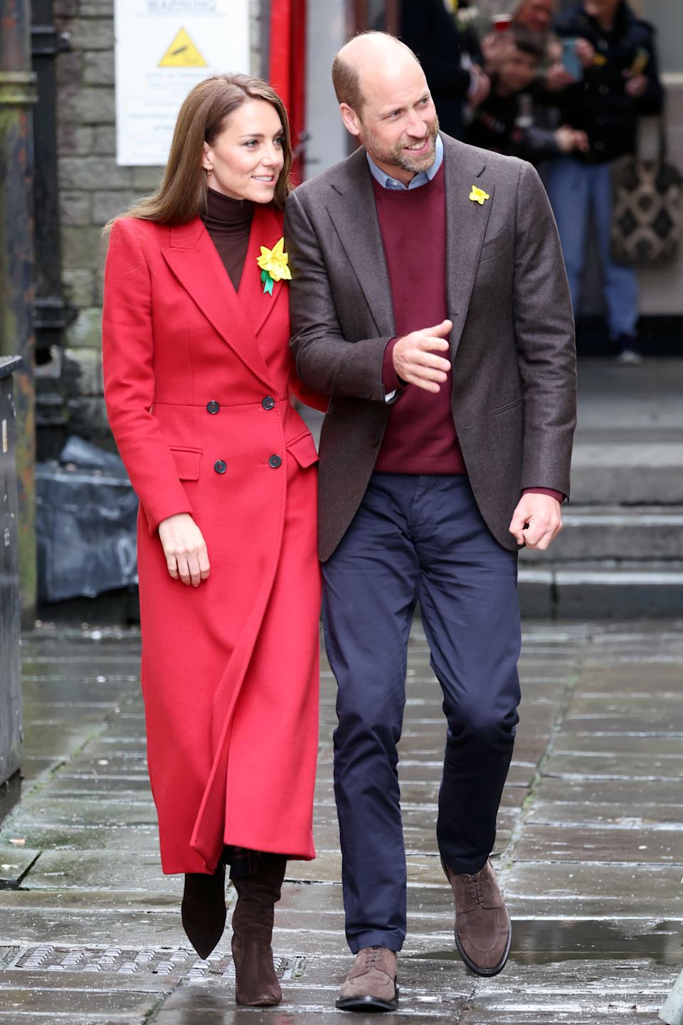 Kate Middleton wearing a red coat and Prince William walking next to her in a blazer and blue pants in Wales smiling and looking to the side