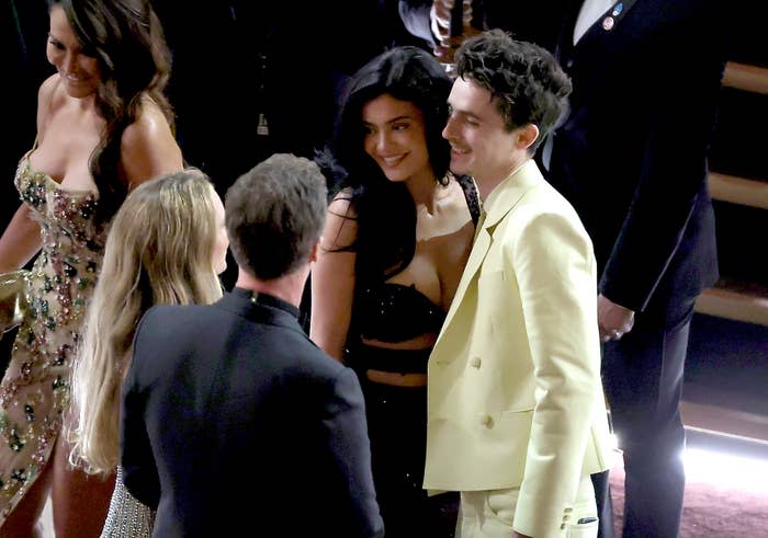 Kylie Jenner and Timothée Chalamet conversing at the Oscars. Kylie wears a strapless black dress; Timothee a yellow suit
