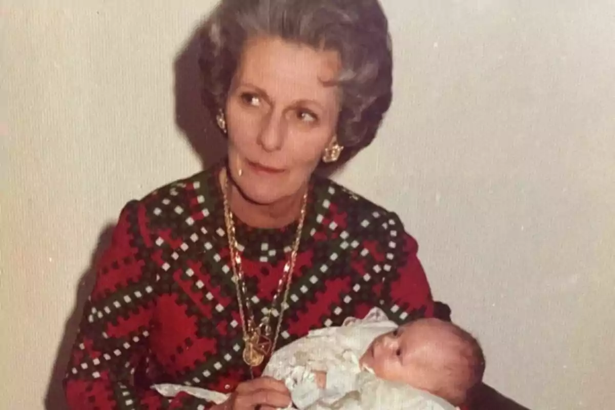 An older woman with gray hair and a colorful dress holds a baby wrapped in a white blanket. An older woman with gray hair and a colorful dress holds a baby wrapped in a white blanket.