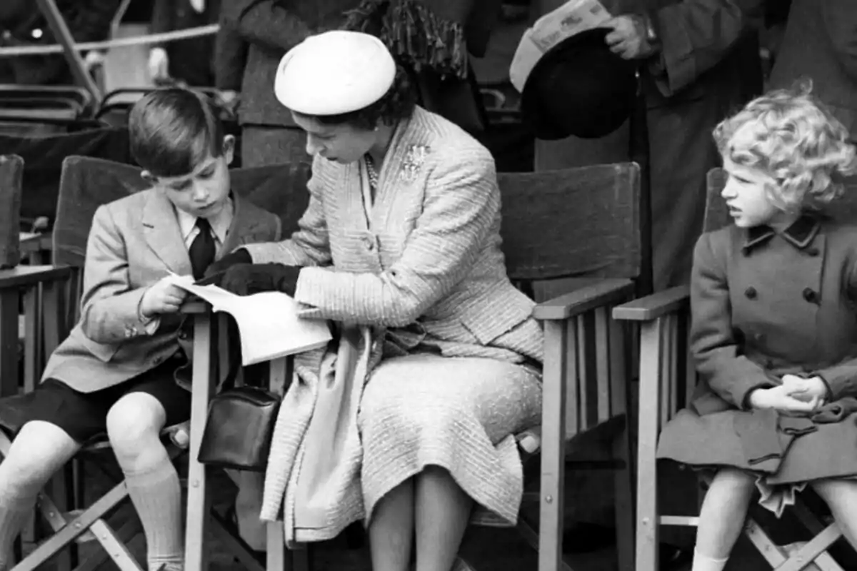 A woman elegantly dressed with a hat and black gloves is sitting between two children on folding chairs, while looking at a book held by one of the children. A woman elegantly dressed with a hat and black gloves is sitting between two children on folding chairs, while looking at a book held by one of the children.