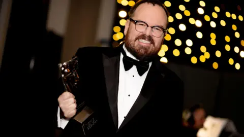 Getty Images Banjo Beale - a bearded man with dark hair and glasses, dressed in a black suit and tie, - holds n award in one had while smiling happily for the camera.