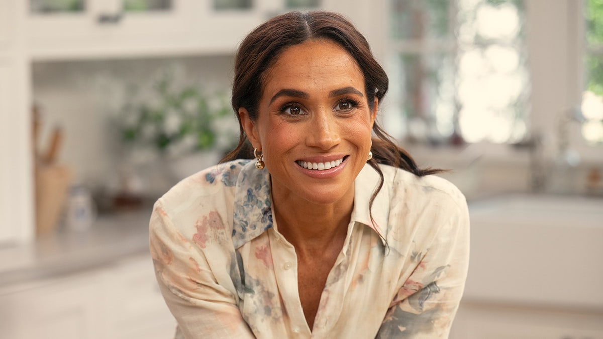A close-up of Meghan Markle smiling and wearing a white and floral shirt dress.