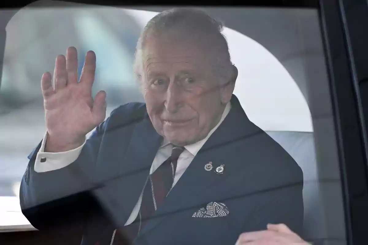 Charles III in a blue suit waves from inside a vehicle.