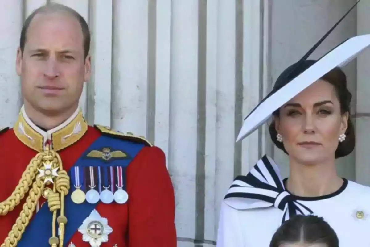 A couple dressed formally, the man wearing a red military uniform with medals and the woman wearing an elegant white dress with a matching hat, both standing in front of a background of columns.