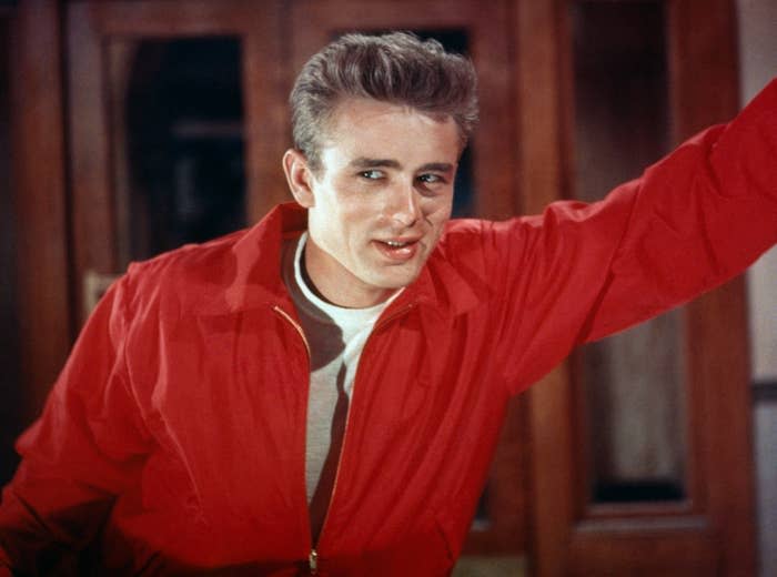 Person posing with one arm raised, wearing a casual jacket over a T-shirt, in front of a blurred background