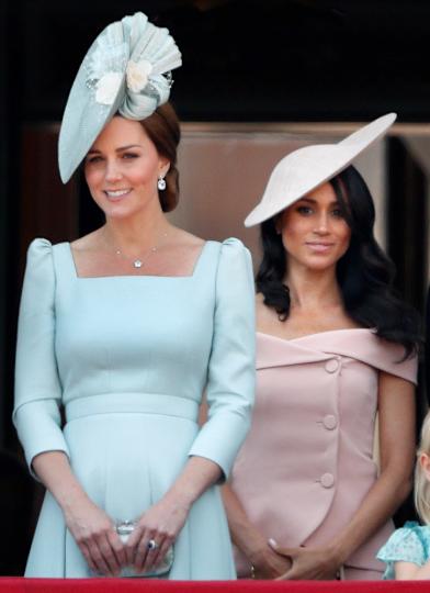 Kate Middleton wearing a light blue dress and a matching hat standing in front of Meghan Markle wearing a powder pink dress and a matching hat.