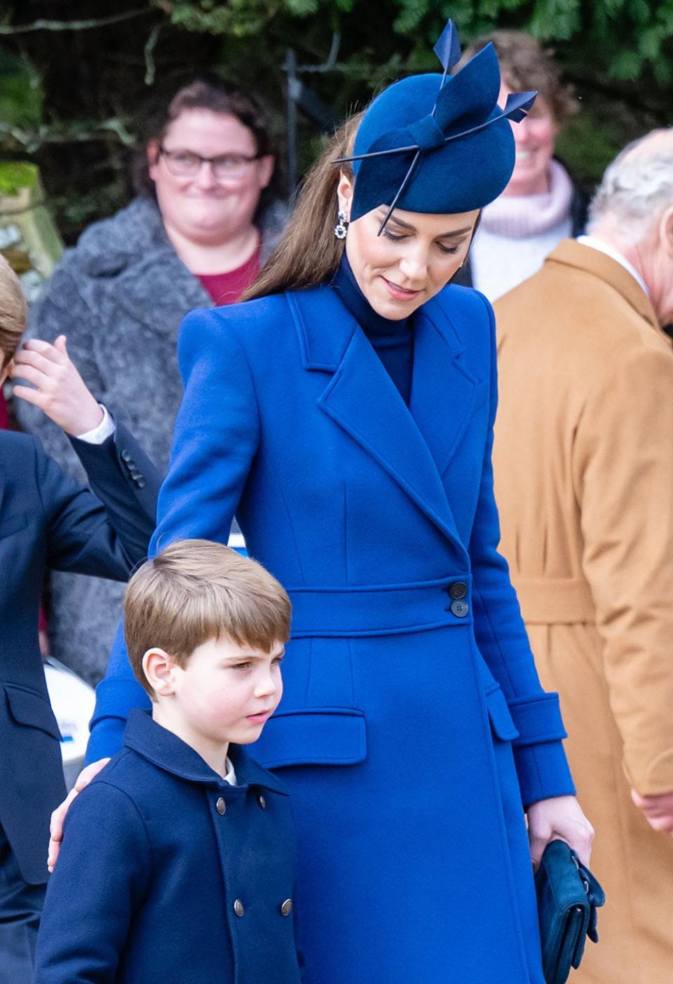 Kate Middleton in a blue coat dress touching Prince Louis' back