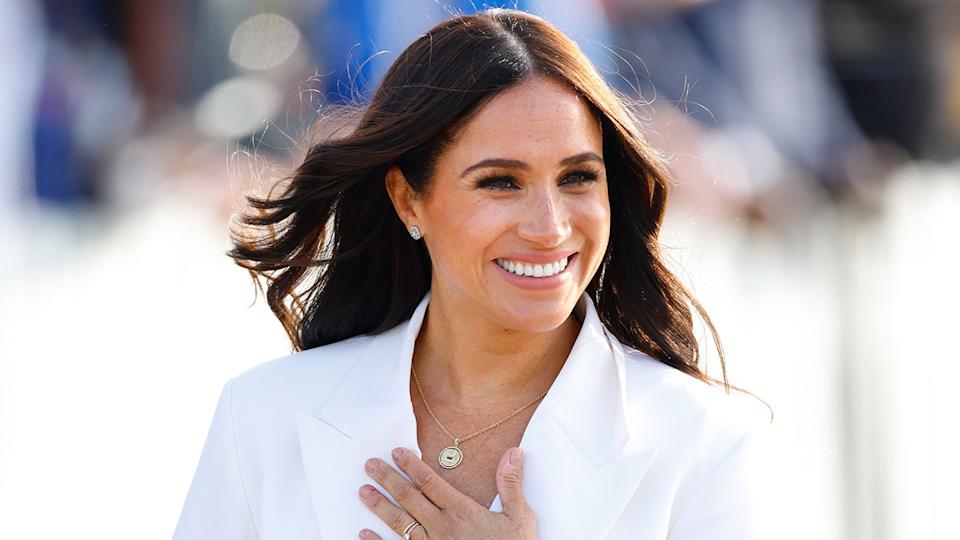 Meghan Markle smiling touching her chest wearing a white blazer.
