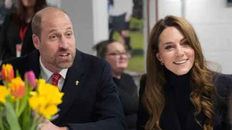 Getty Images William and Kate sitting next to each other and smiling while talking to someone off camera - William has a dark suit and red tie on, while Kate has a dark blue jacket on. Flowers sit in front of the couple.