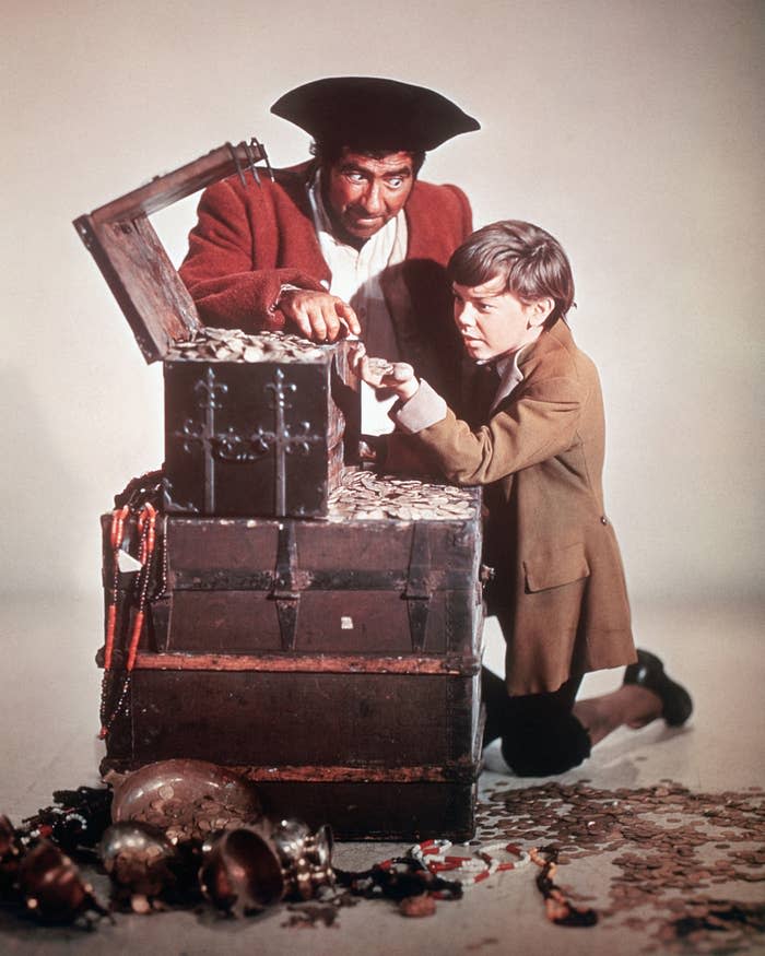 Two people dressed as pirates examine coins in an overflowing treasure chest, with scattered jewels and coins on the ground