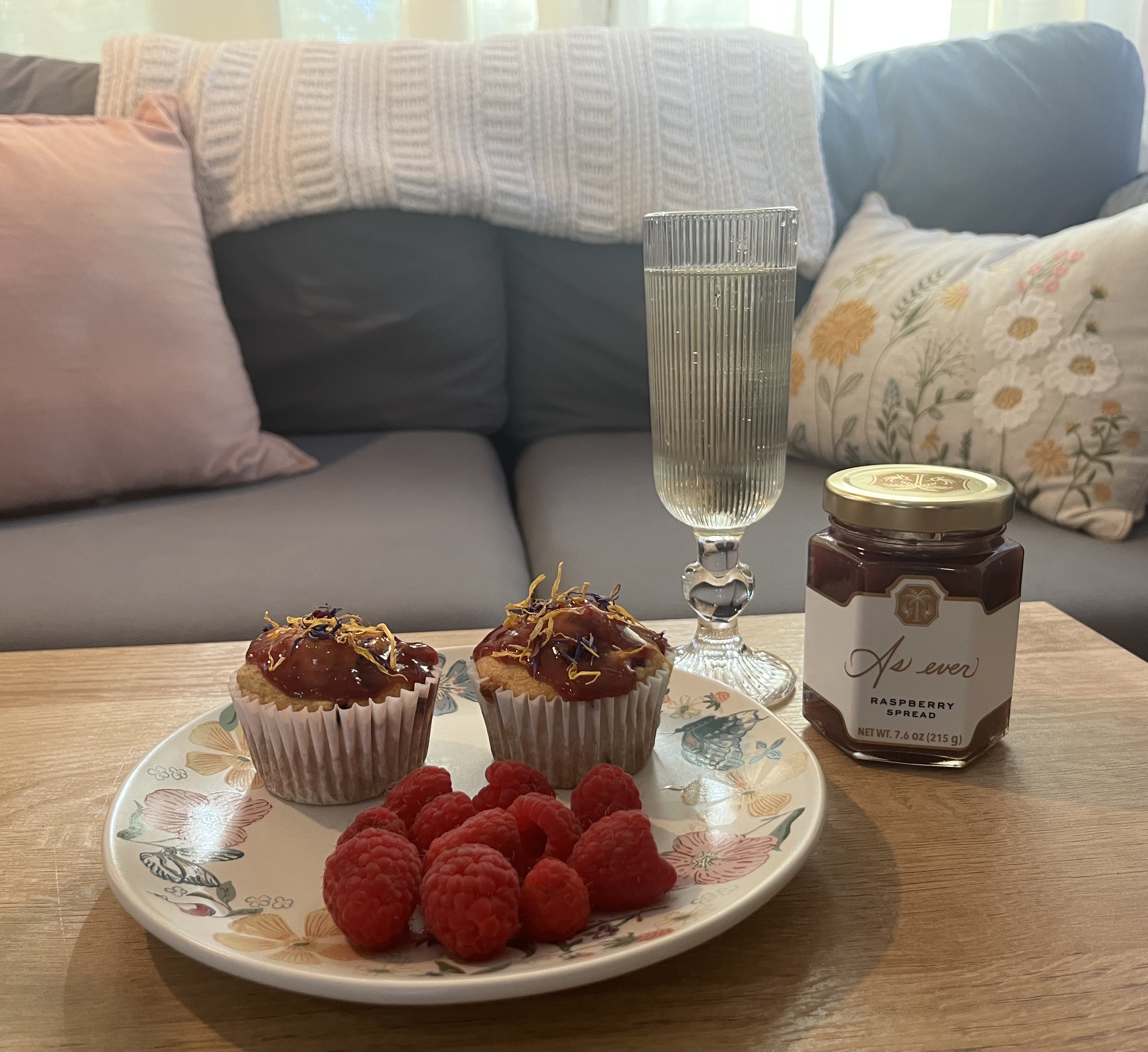 A table with a glass of champagne and a plate of muffins with raspberries on them next to a jar of Meghan Markle's As ever jam.