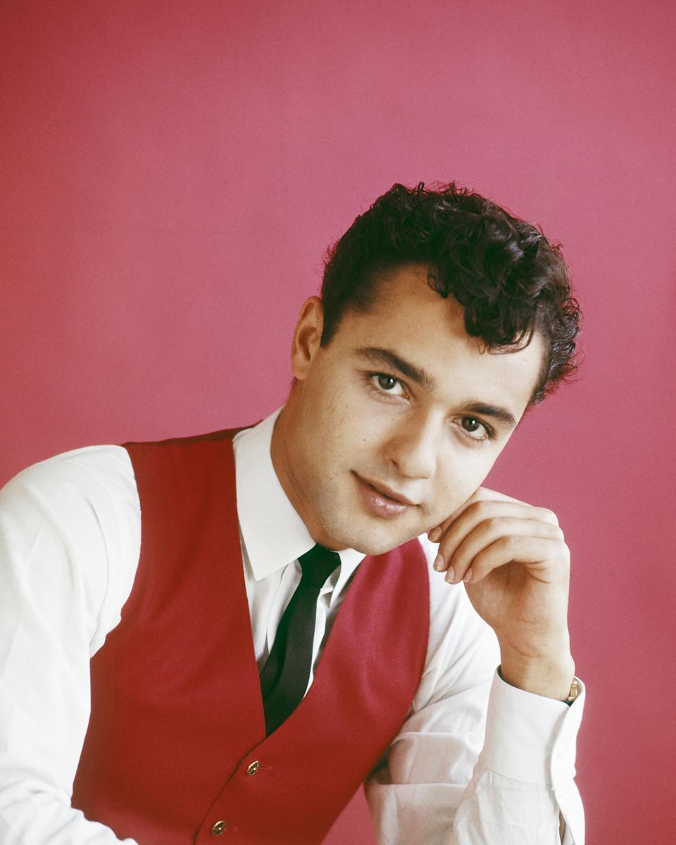 Person wearing a white shirt, red vest, and black tie, posing with hand on chin against a plain backdrop