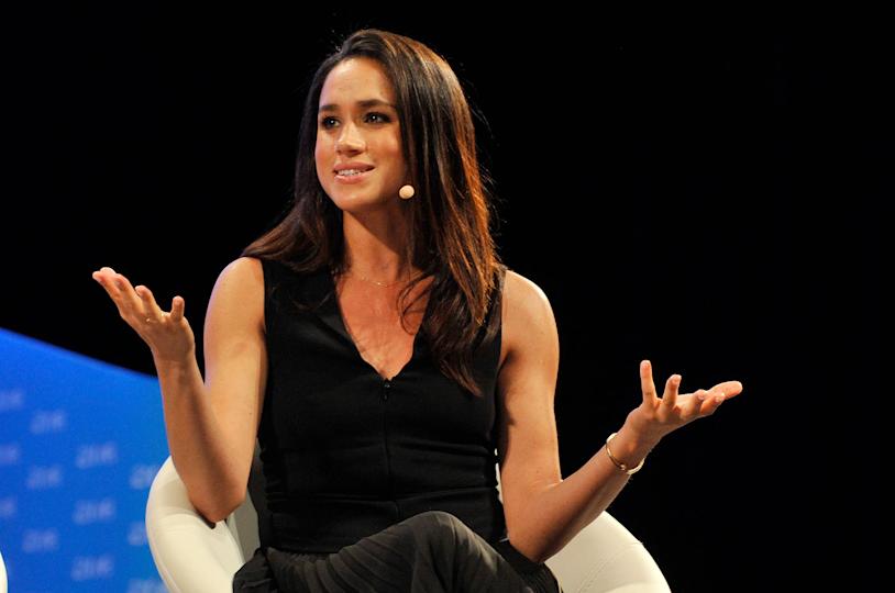 Meghan Markle with her arms raised wearing a sleeveless black dress and speaking to a mic.
