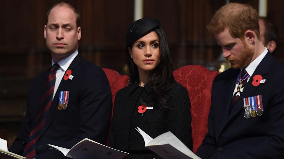 Prince William sitting next to Meghan Markle and Prince Harry in burgundy chairs.