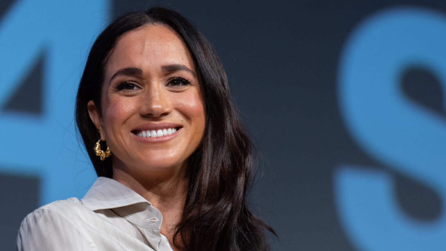 A headshot of Meghan Markle wearing a tan blouse and gold hoop earrings smiling in front of a blue background