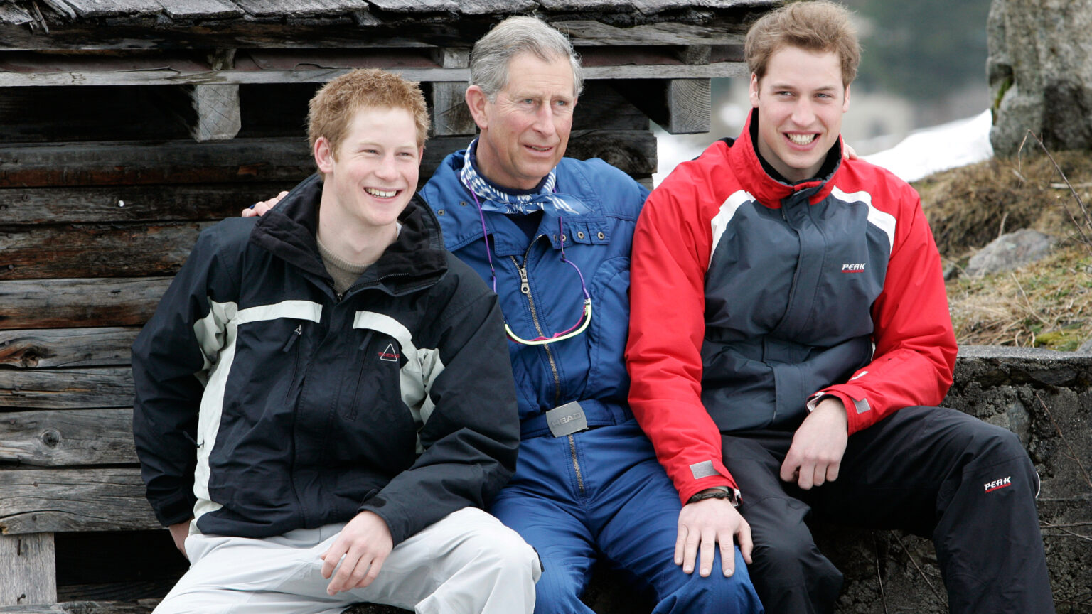 Prince Harry, King Charles, and Prince William wearing ski clothes sitting next to each other smiling in 2005