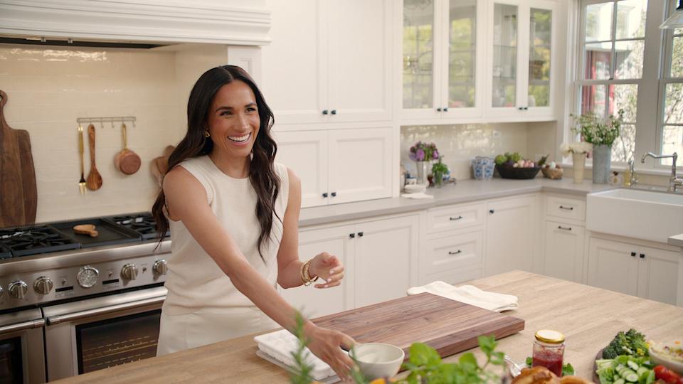 Meghan Markle cooking in a kitchen and smiling wearing a sleeveless ivory top in her show With Love, Meghan