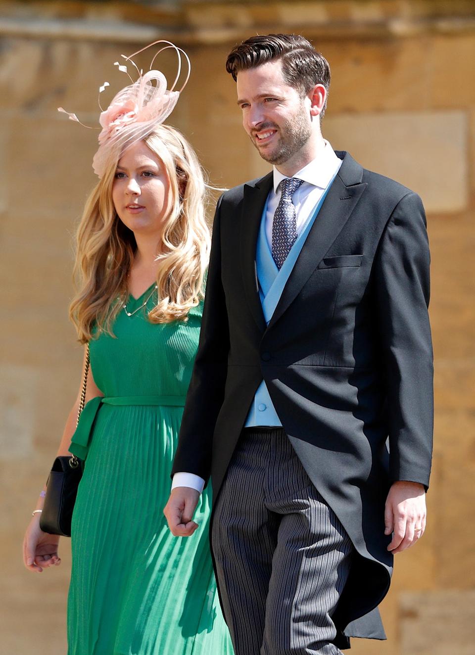 Jason Knauf wearing a suit and tie walking next to a woman wearing a bright green dress and a pink fascinator.