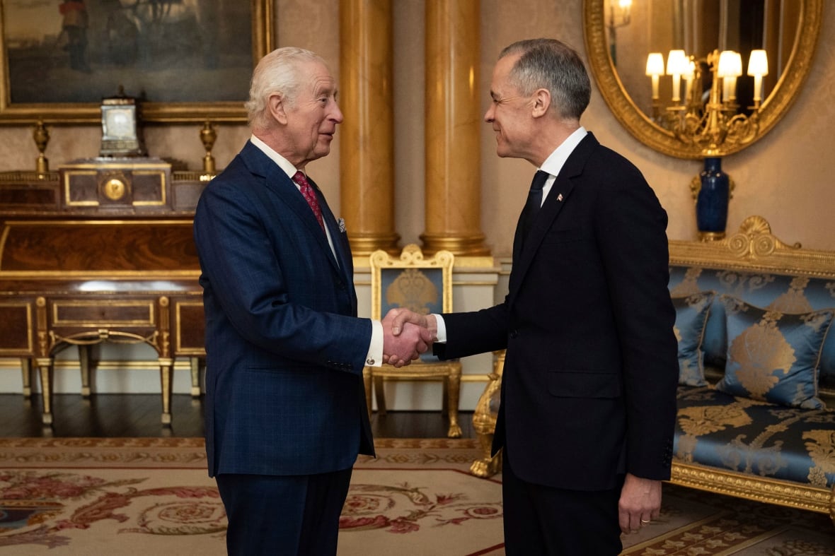 Two people shake hands standing in the centre of a room with furniture against the wall and lights reflected in a mirror.