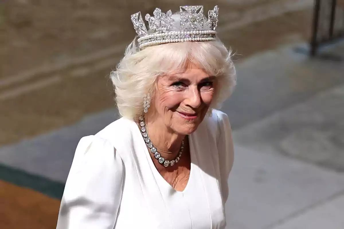 Queen Camilla with blonde hair and a tiara smiles while wearing an elegant white dress.