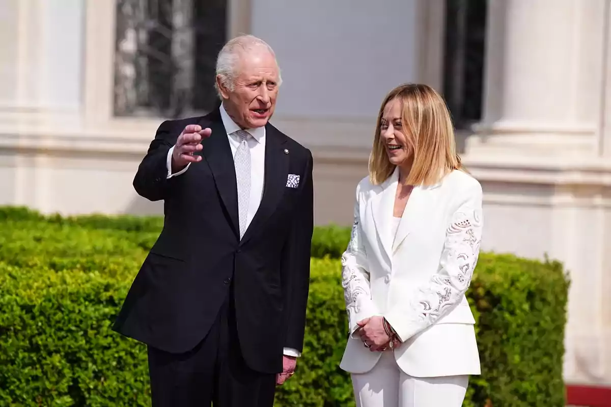 Carlos III and Giorgia Meloni, dressed formally, converse outdoors in front of an elegant building with green shrubs. Carlos III and Giorgia Meloni, dressed formally, converse outdoors in front of an elegant building with green shrubs.