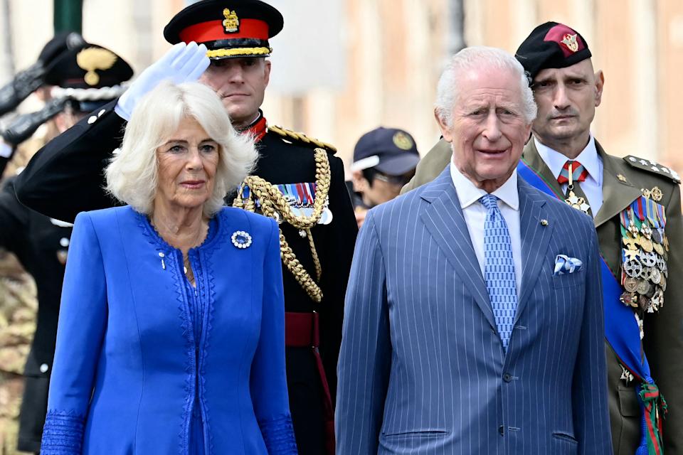 Queen Camilla wearing a blue coat with a diamond brooch standing next to The King and smiling standing in front of soldiers on April 8, 2025