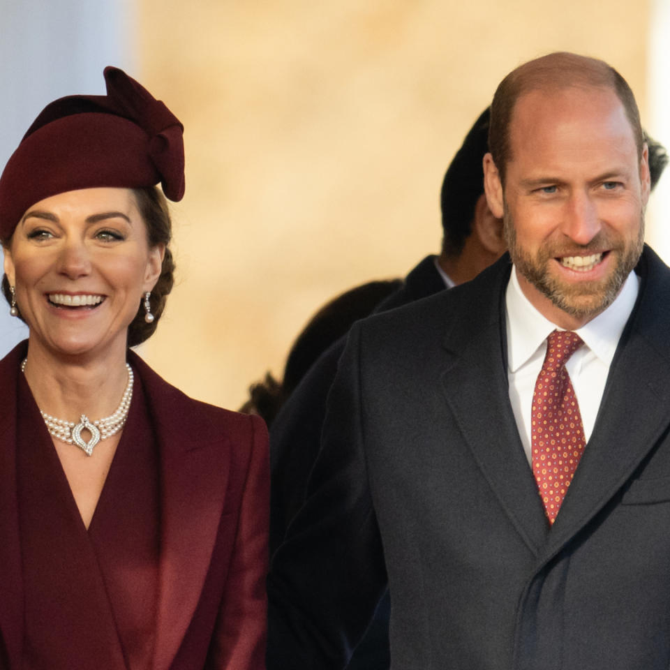 Kate Middleton wearing a maroon coat and hat and smiling next to Prince William wearing a dark coat and a red tie .