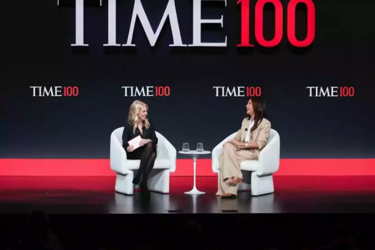 Two people sitting on white chairs converse on a stage with the 