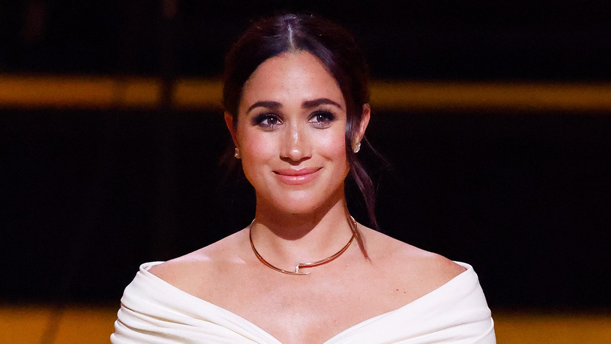 Meghan Markle smiling wearing a white stylish top and a gold necklace.