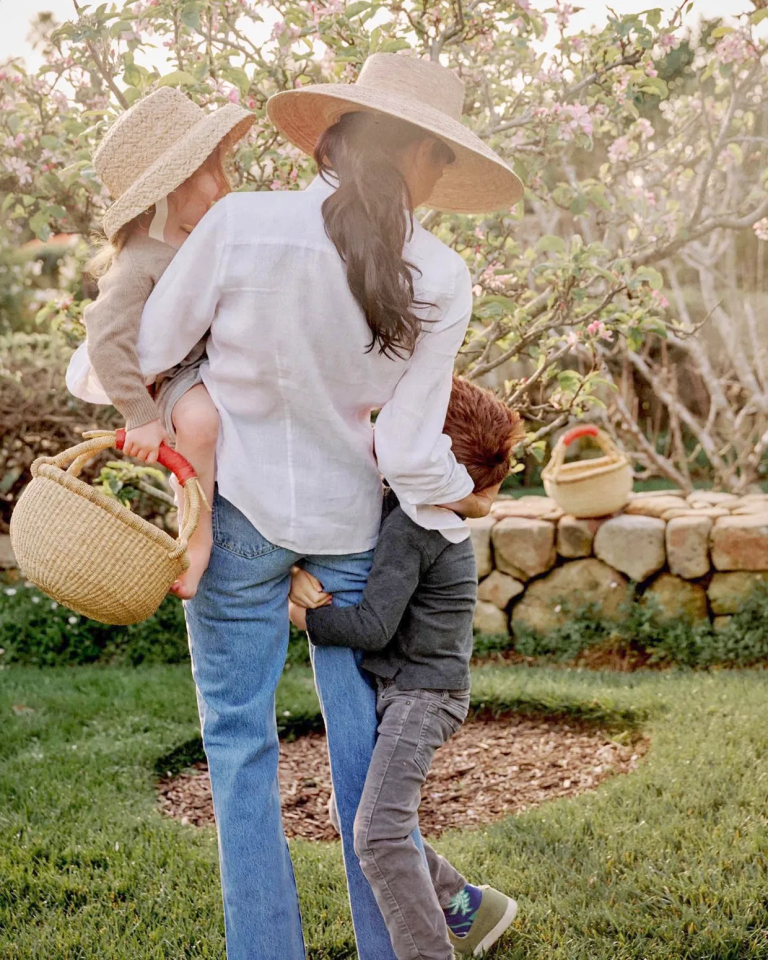 Woman carrying a child while another child holds onto her legs in a garden.
